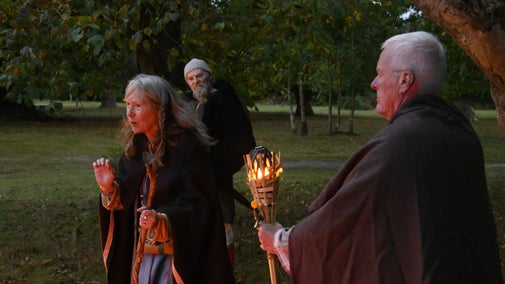 Three people in Anglo-Saxon dress, one holding a lit torch while another is telling a story